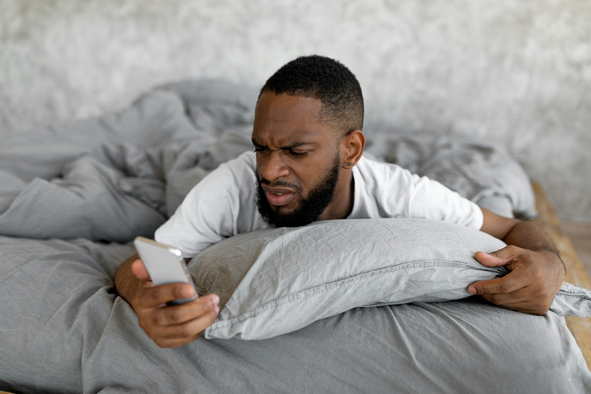 Confused black man looking at phone in bed